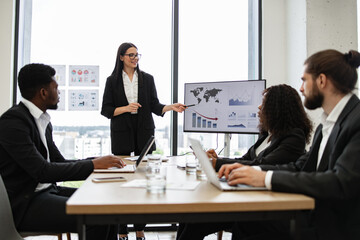 Business woman gives report to multiracial colleagues standing near graphs on monitor. Professional team discussing data analysis, growth charts, and performance metrics in modern office setting.