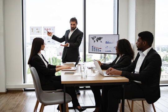 Bearded business man giving presentation to multiracial colleagues standing near graphs and charts on window glass. Professional businesspeople collaborating, discussing ideas and growth strategies