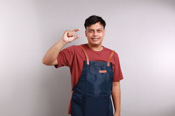 Asian young man wearing apron over white background smiling and confident gesturing with hand doing small size sign with fingers looking and the camera. measure concept.