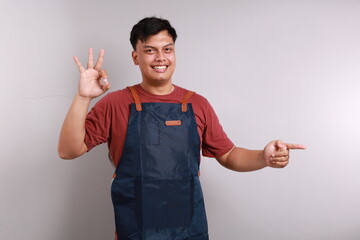 Young asian man barista barman employee wear blue apron work in coffee shop pointing sideways while making okay hand gesture sign