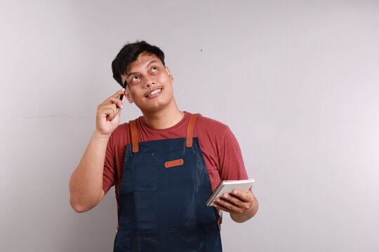 Asian barista man in blue apron taking order, writing on menu book list, thinking with pen on head. Isolated image on white background