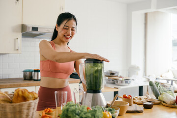 Woman in Sportswear Preparing a Healthy Green Smoothie in a Modern Kitchen with Fresh Vegetables...