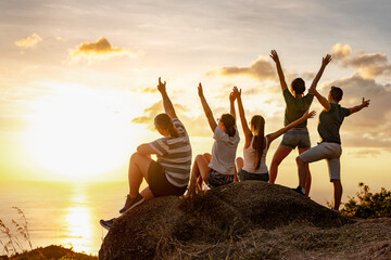 Group of happy friends are sitting at big rock at mountain top and enjoys sunset