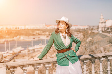 Obraz premium Woman walks around the city, lifestyle. Happy woman in a green jacket, white skirt and hat is sitting on a white fence with balusters overlooking the sea bay and the city.