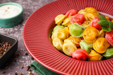 Plate of boiled colorful dumplings with basil and bowl of sour cream on dark table