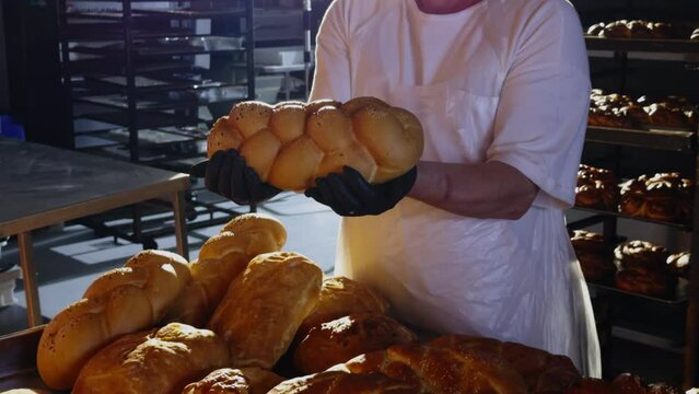 The baker is holding a braided bun taken out of the oven. Image from a bakery with several baked dough products taken out of the oven. Dough products ready for sale. Successful small business