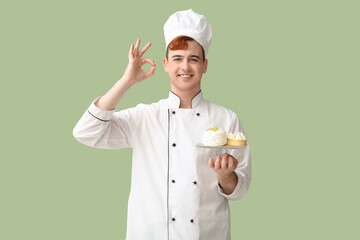 Male confectioner in uniform holding stand with tasty desserts and showing OK gesture on green background