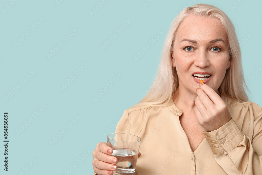 Wall mural Beautiful mature woman with glass of water taking pill on blue background