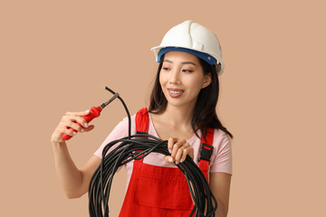 Young female electrician with pliers and cables on beige background