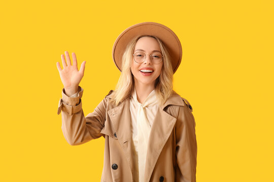 Beautiful young stylish woman waving hand on yellow background