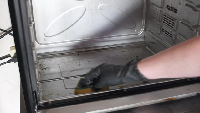woman's hand in a black glove washes the oven with sponge and removes black burnt drops of fat from the oven, kitchen cleaning, routine, High quality 4k footage