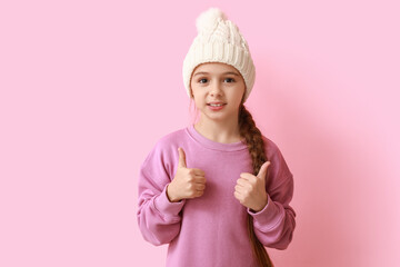 Cute little girl in warm hat showing thumbs-up on blue background