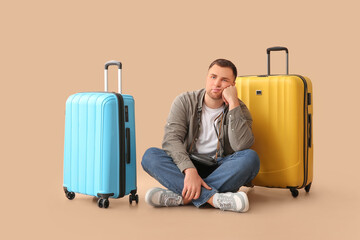 Handsome young bored male tourist with suitcases sitting on beige background