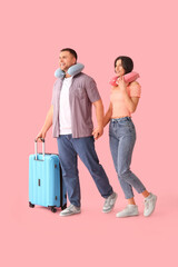 Beautiful young happy couple of tourists with suitcase and travel pillows on pink background