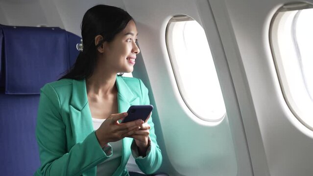 A young Asian businesswoman, dressed in a formal suit, sits at her airplane seat with a laptop and briefcase, utilizing in-flight Wi-Fi for a conference call.