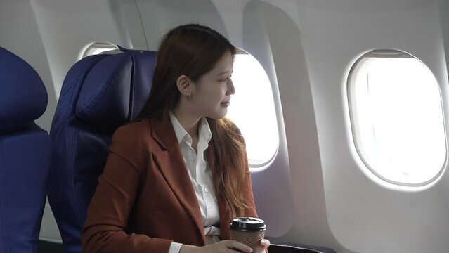 A young Asian businesswoman, dressed in a formal suit, sits at her airplane seat with a laptop and briefcase, utilizing in-flight Wi-Fi for a conference call.