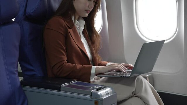 A young Asian businesswoman, dressed in a formal suit, sits at her airplane seat with a laptop and briefcase, utilizing in-flight Wi-Fi for a conference call.