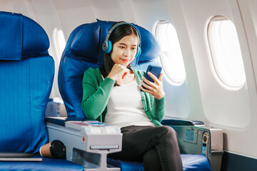 A young Asian woman, an airplane passenger, relaxes by listening to music on her mobile phone. She uses technology to unwind during the long journey.