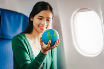 A young Asian woman, an airplane passenger, sits at her seat holding a world globe. Her travel essentials include a boarding pass, Earth day, Sustainable Fuel and Green energy, Save the world.