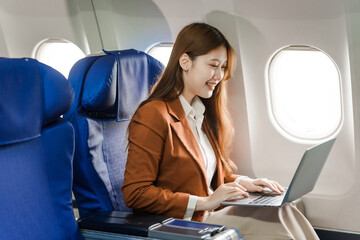 A young Asian businesswoman, dressed in a formal suit, sits at her airplane seat with a laptop and briefcase, utilizing in-flight Wi-Fi for a conference call.