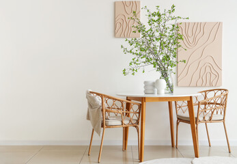 Table, armchairs and vase with blossoming branches near white wall in beautiful dining room