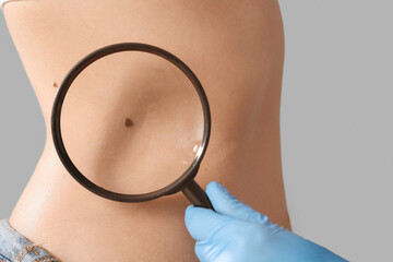 Dermatologist with magnifier examining mole on woman's belly against light background, closeup