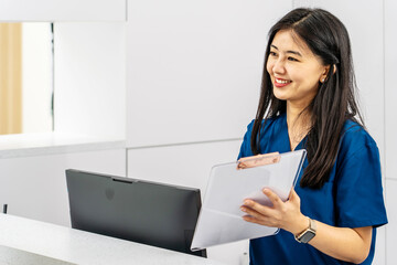 Asian female administrator talks with patient. Adult woman stands near reception desk in clinic...