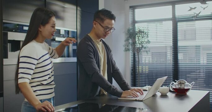 Nice sunny morning. Busy Asian male working hard on his laptop. Entering data. Beloved wife or girlfriend coming up to man. Husband pouring some hot tea for woman. Spending weekend together.