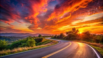 Naklejka premium Vibrant orange and pink hues illuminate the sky as the sun sets behind a winding asphalt road, leaving ample copy space for text or design.