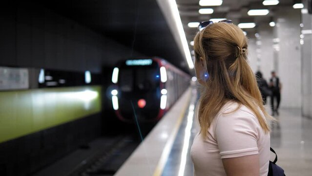 Side view of a young woman with a backpack at the metro station, the train is moving along the rails. A woman in the subway is waiting for a train.
