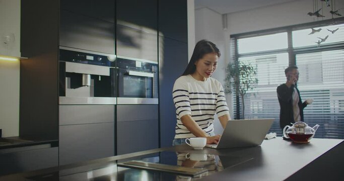 Camera sliding around pretty Asian woman. Young girl working on laptop. Busy man speaking on smartphone and sipping coffee or tea in background. People freelancing from home in kitchen.