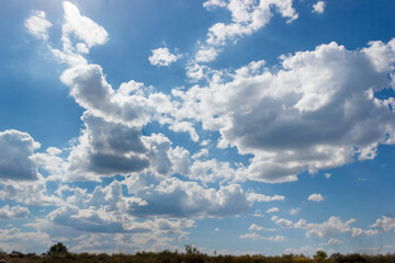 A natural landscape with azure sky, fluffy cumulus clouds, and the sun shining through, creating a picturesque scene over the grassy meadow and horizon.