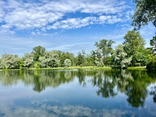 summer lake in the park, blue sky with white clouds and green trees reflection on the lake surface