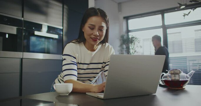 Beautiful Asian woman looking at her laptop screen. Tea pot standing next to laptop. Woman pressing down keys. Entering information. Husband or boyfriend walking around while talking on phone.
