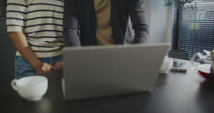 Asian couple standing close to each other. Launching computer. Opening lid of laptop. Waving their hands to camera. Remote communication with friends or relatives. Relaxing in kitchen.