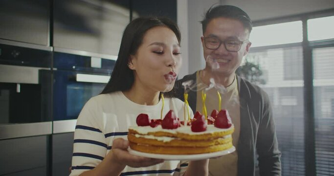 Warm moment. Asian couple celebrating birthday together. Woman blowing out candles on her birthday cake. Man hugging girl from behind. Preparing for memorable evening. Celebration.