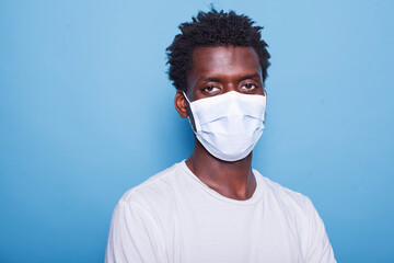Close-up of black guy with face mask looking at camera, in front of isolated blue background. Portrait of African American man with afro hair, having protection against covid 19.
