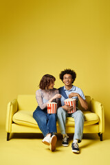 Obraz premium A man and a woman are seated together on a yellow couch, sharing a loving embrace in a sunlit room.