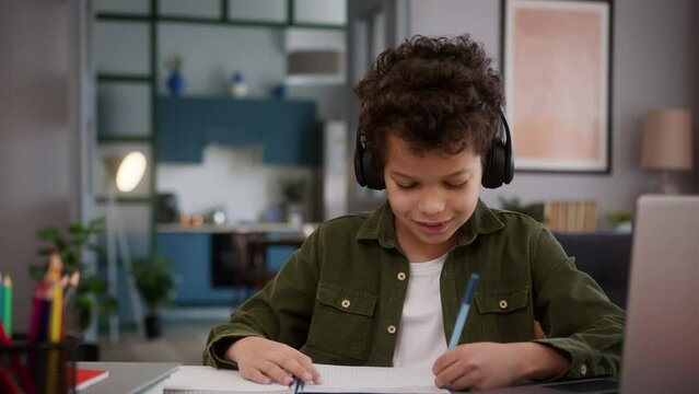 Hispanic African American little boy child kid son pupil schoolboy funny happy at home distant education e-learning studying at table writing task write homework listening music in headphones dancing