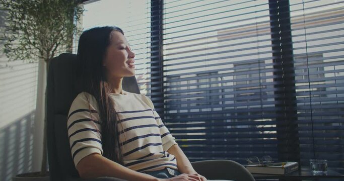 Zooming in on charming Asian woman sitting on chair in modern office room or flat with panoramic window. Dreaming about something. Joyfully smiling while glancing at camera. Wellbeing. Leisure concept