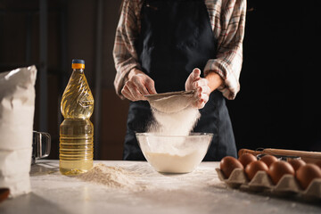 Close-up of woman sifting flour through sieve into transparent bowl while baking at home. Flour dust is visible in the air, dynamic scene.