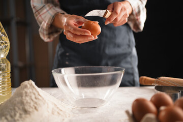 Close-up of female hands cracking an egg with a knife over a clear bowl. Flour, eggs, and a bottle of cooking oil are on the table. Preparing dough for baking in a home kitchen