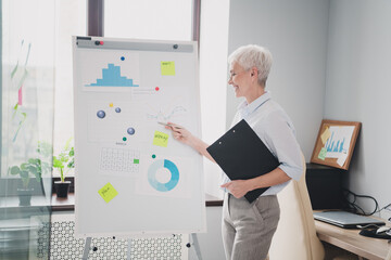 Photo of smiling positive mature lady specialist formalwear analyzing graphs indoors workplace...