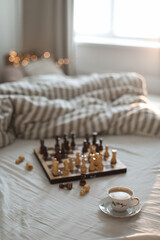 Chess board with cup of coffee on bed. Strategy thinking. Morning ideas concept. Pastel shades. Top view of unmade bedding sheets and blanket