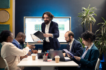 Executive team develops strategic plans and financial projections during meeting, manager provides clipboards with data reports. Employees allocating essential resources from capital funds.