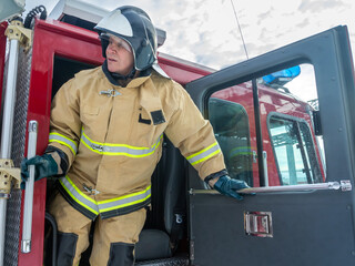 A firefighter in a helmet and protective clothing opened the door of a fire truck.  A firefighter gets out of a fire truck to start extinguishing the fire. The rescue service.