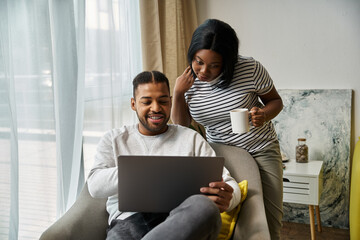 Fototapeta premium African American couple cozy at home, using laptop and enjoying drink.