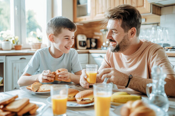 A father and son are enjoying breakfast together, with the boy eating sandwiches while his dad sips orange juice in their bright kitchen.
