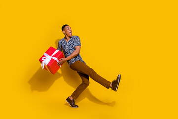Fototapeta na wymiar Full length photo of positive guy dressed print shirt preparing valentine day gift walking empty space isolated yellow color background
