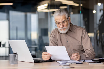 Senior businessman reviewing documents while sitting at a desk with a laptop in a modern office setting.